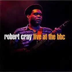 Picture of Cray's upper body while he's playing guitar against a dark background. He is wearing a purple and black horizontally-striped shirt and appears to be wearing a black beret. The words "robert cray live at the bbc" appear in lowercase font with "robert cray" in white and the rest in orange.