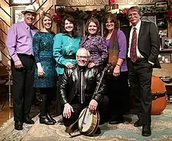 Seven white adults in semi-formal clothing are standing on a stage in front of a wooden sign that reads "Carter Fold".