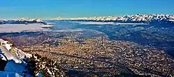 Image of a city viewed aerially, looking down from a mountain with some faint clouds hanging above the cityscape. Other mountains, Chartreuse, Mont Blanc, and Belledonne visible in the background.