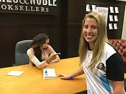 A young dark-haired woman sits at a desk in a bookstore, pointing to a blonde woman wearing her likeness on a t-shirt.