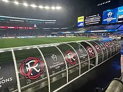 A covered bench with a New England Revolution and CONCACAF Champions Cup logo in front of a soccer field at an empty soccer stadium at night.