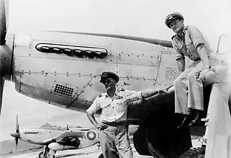 Two men in military uniforms and peaked caps, with a single-engined fighter plane