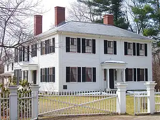 A white two story house, whose main section is square, although there is a wing extending off the back. There is a white fence with a gate across a gravel drive.