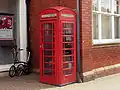 A red, cast iron telephone kiosk with a domed roof. it stands against a red brick wall at the corner of a building.