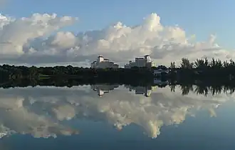Across a body of water is land covered with trees and with two large. In the middle are two large buildings that look like hotels. The water is calm and reflecting the blue sky and puffy white clouds above.