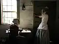 Living History interpreters portraying two young women of Salem. The seated woman is copying Bible verses using a quill.