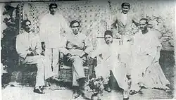 Six men: four seated and two standing