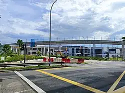 The entrance to the SRTC with a fenced elevated track running across the background. The foreground includes a road intersection marked with yellow box junction lines and pedestrian crossings.