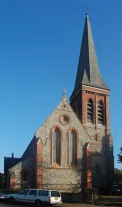 Side view of a long flint-built church with several parts, each with a different roofline. A spire is visible behind the body of the church. All windows are framed by brickwork.