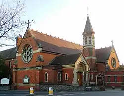 Three-quarter view of an ornate brick building with pale stone dressings and recessed lancet and round windows. A rose window dominates the left-hand side. Perpendicular to its right, there is an arched entrance porch; beyond that is an octagonal tower with louvre-style openings below a conical spire.