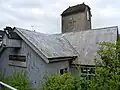 Very close view of a low iron church with a corrugated iron roof and a stone tower. This has a low brown cap-style roof and a projecting crucifix.