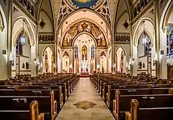 Interior view from the rear of the center aisle of a large, well lit, neo-Gothic church, toward the colorful apse.
