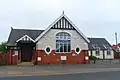 A cottage-style building with a modern extension to the left. The main body is of brick and white-painted pebbledashing; there is some timber-framing below the roofline. There are two round windows and two white plaques with the words "GOD IS LIGHT" in ornate writing. These flank a wide arched window.